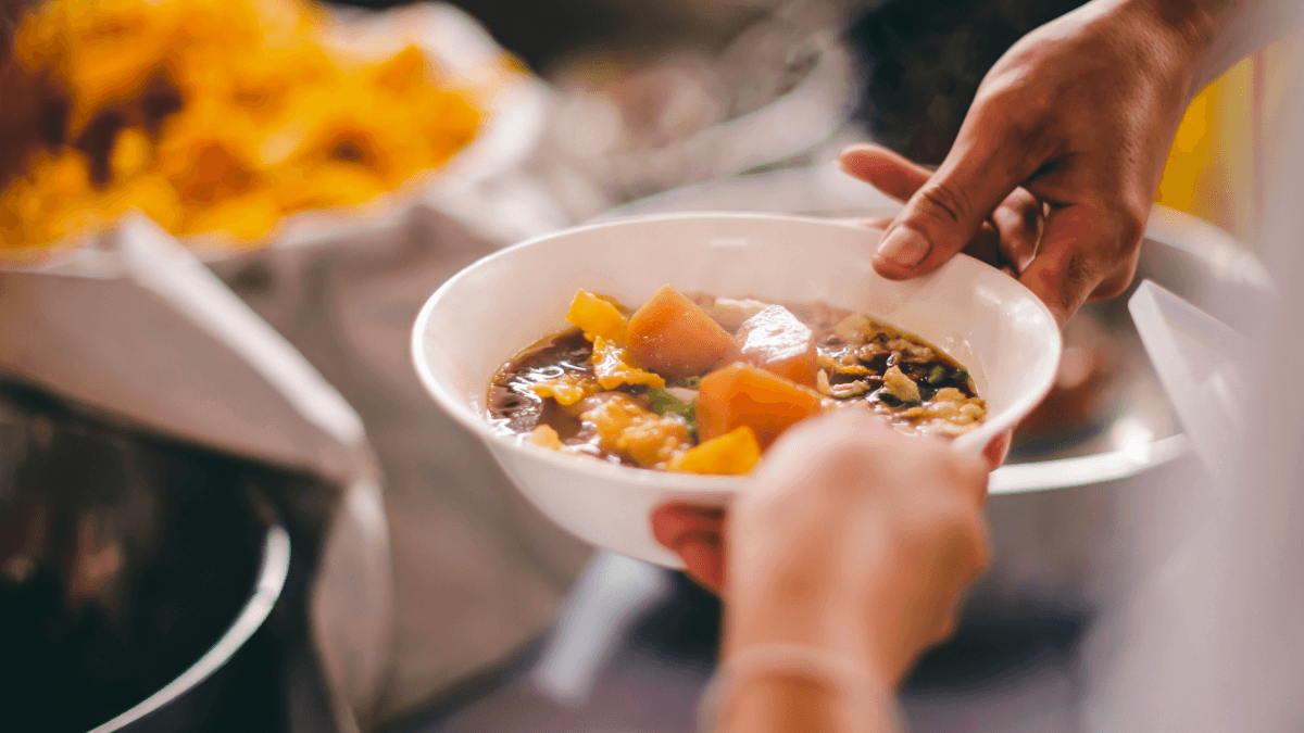 Person receiving a bowl of soup from another person. The soup bowl is white and has large pieces of orange vegetables in it.