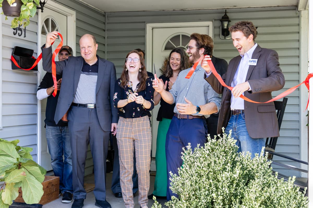 Group of seven people standing on a porch in front of a home, holding a cut red ribbon. They are celebrating and are mid-clap.