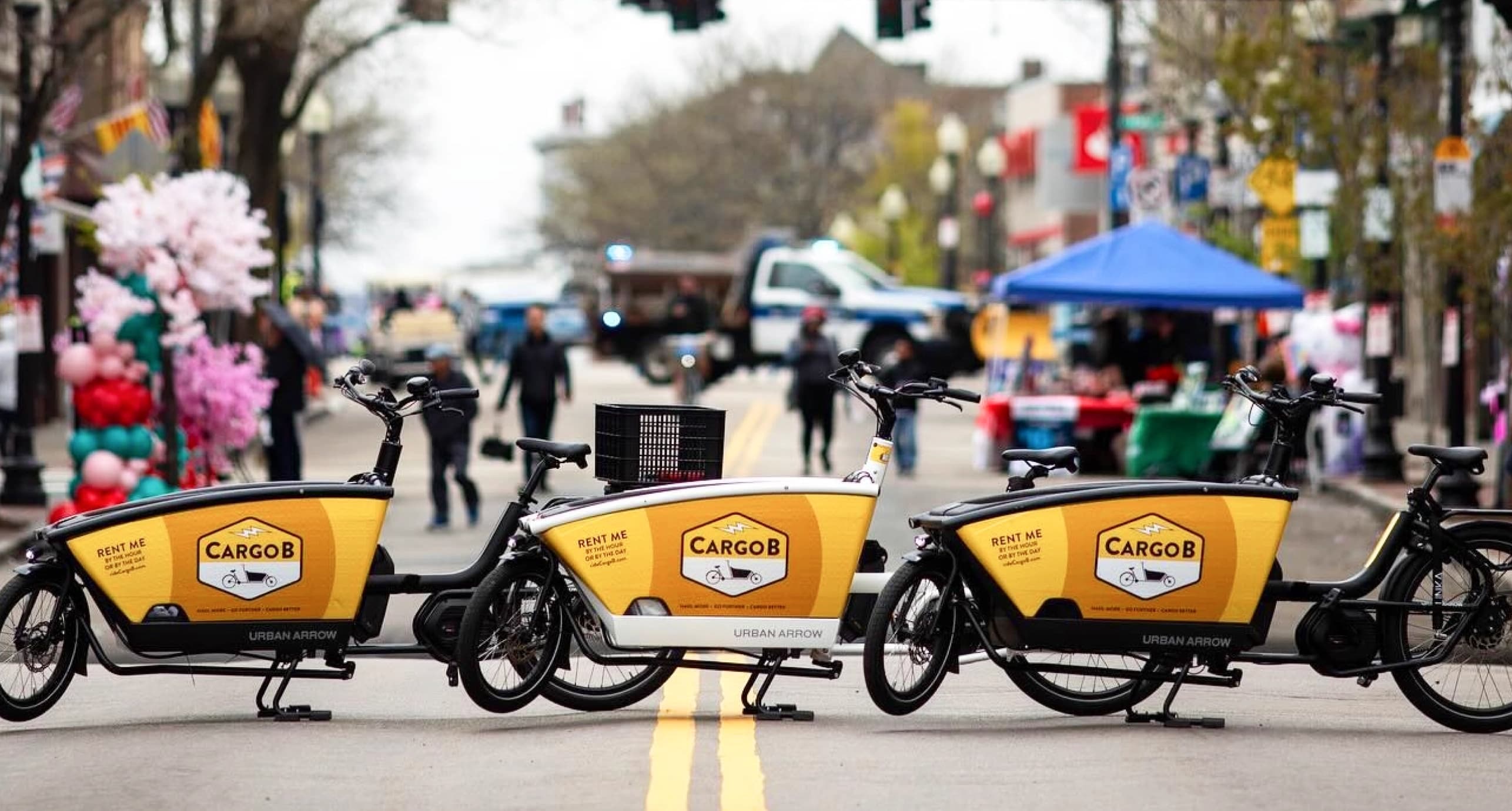 Three cargo e-bikes with yellow boxes are parked on a street during an outdoor event, with tents and people in the background.