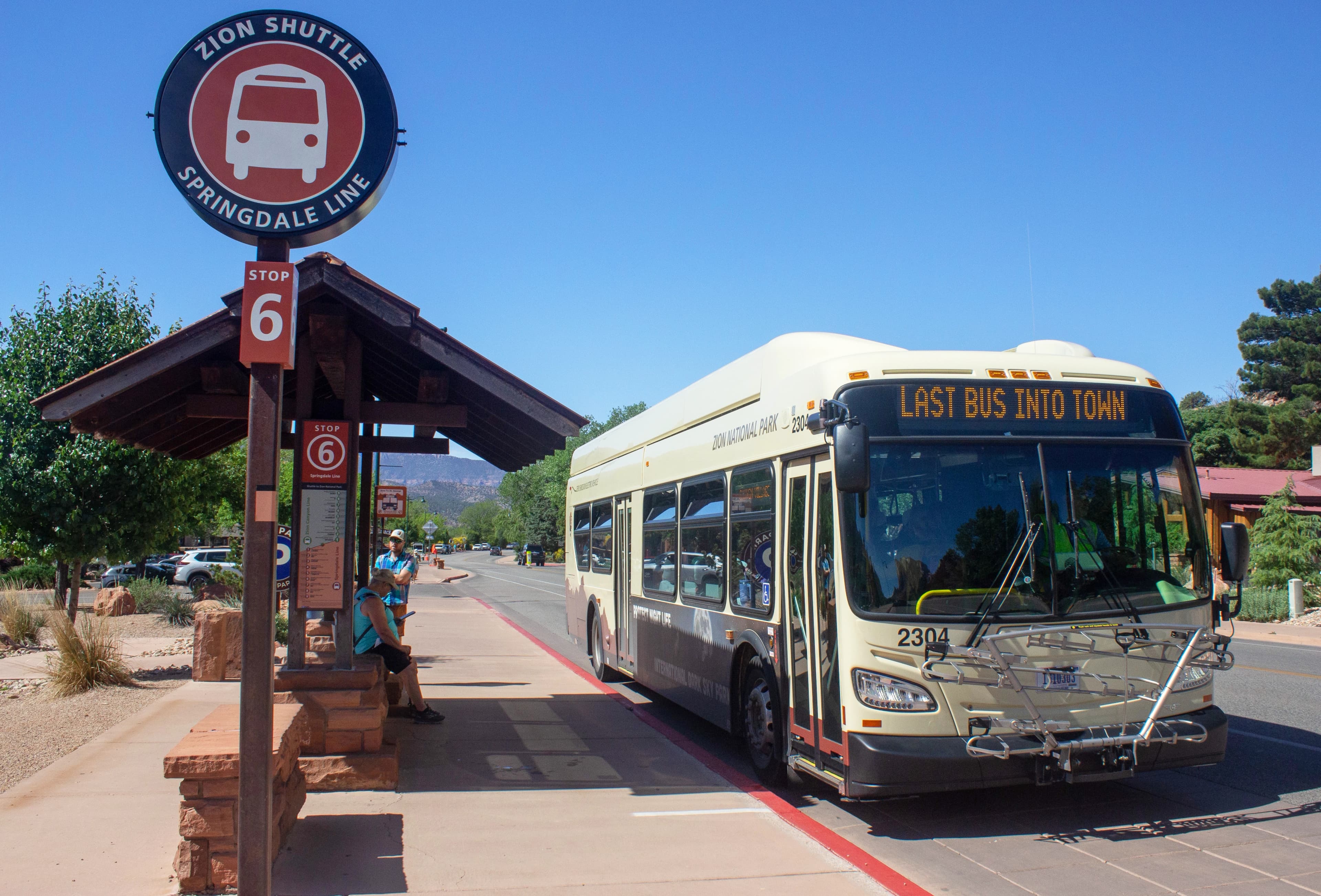 A Zion electric shuttle bus labeled "Last Bus Into Town" is parked at Stop 6 in Springdale, with a few people nearby under a wooden shelter.