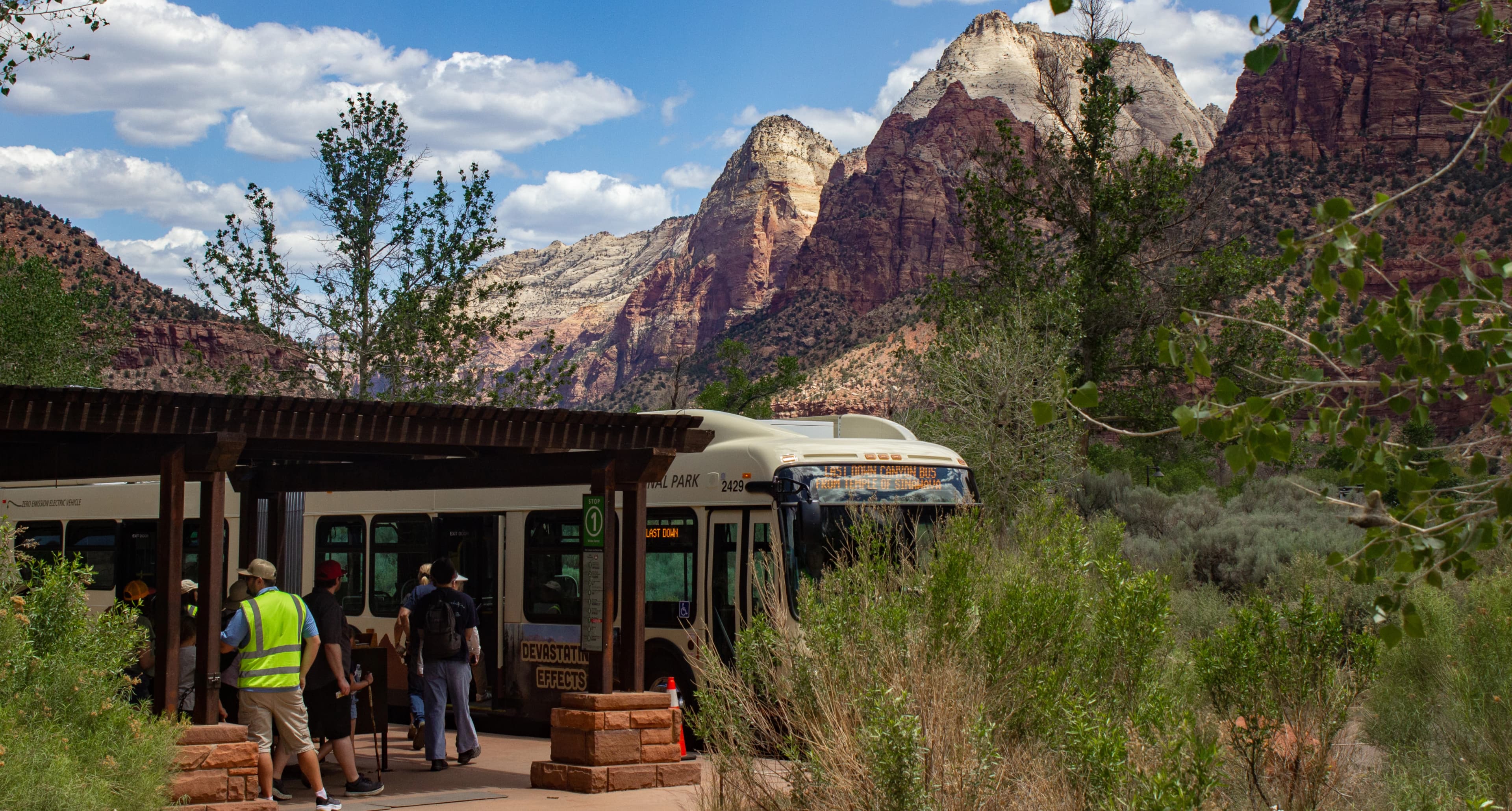 People boarding a parked electric shuttle bus at a scenic stop in Zion National Park, with towering red rock formations and trees in the background.