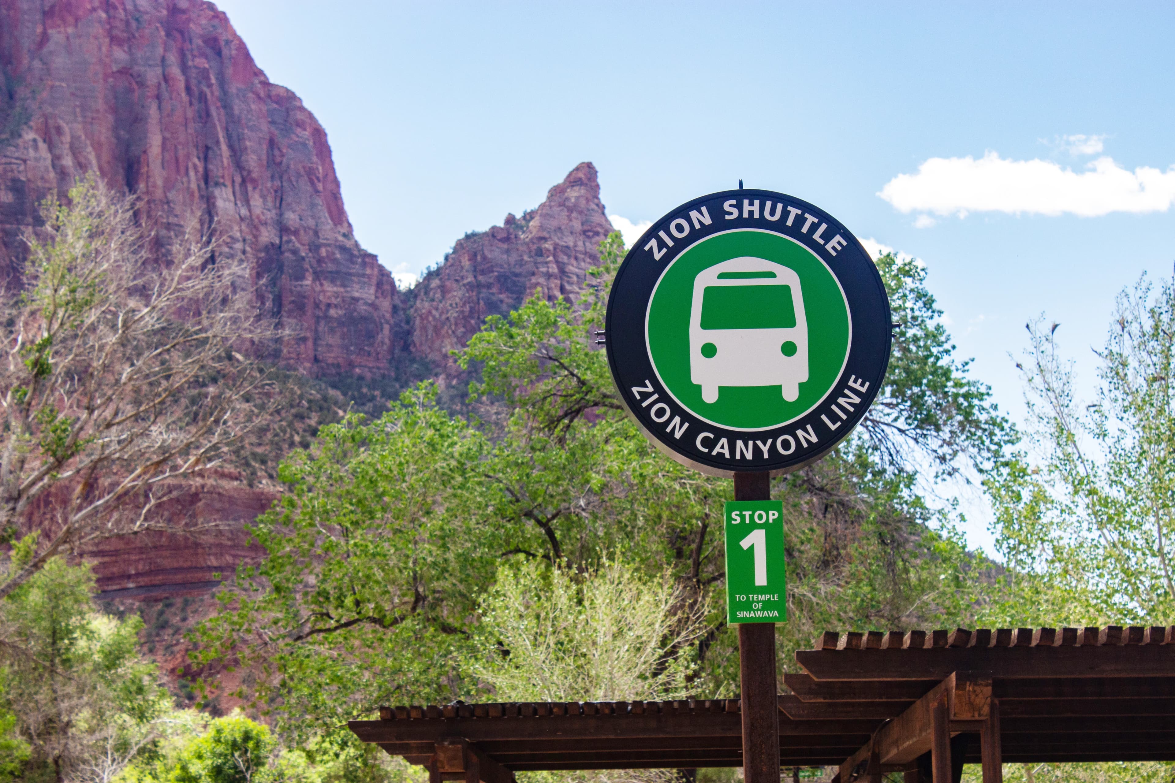 Sign for Zion Shuttle, Stop 1, against a backdrop of red rock cliffs and green trees under a blue sky.