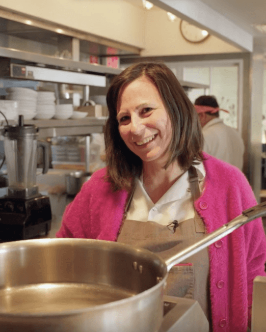 Photo of a woman in a restaurant kitchen looking at the camera, with short brown hair, a ping sweater, a linen apron and a smile.