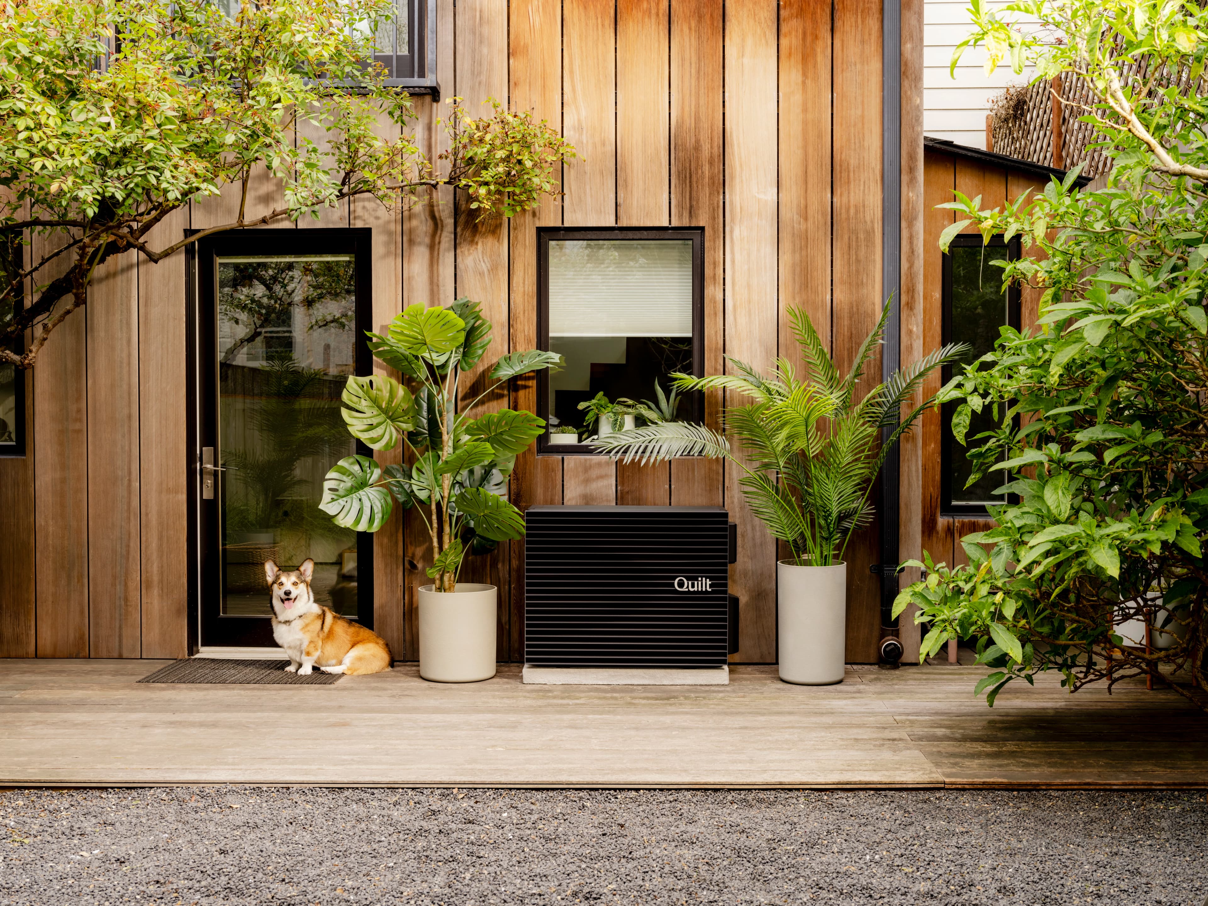 A modern, energy-efficient heat pump unit labeled "Quilt" is installed outside a stylish wooden-clad home. The unit is surrounded by lush green plants in sleek, neutral-colored pots. A corgi sits on a doormat near a glass door, looking at the camera. The scene is framed by natural elements, including trees and bushes, enhancing the warm and inviting ambiance of the outdoor space.